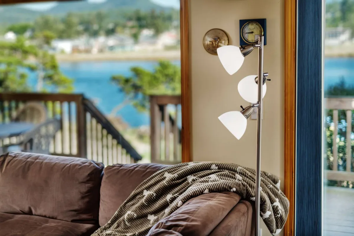 A comfortable interior scene with a sofa positioned to enjoy tranquil water views through a large window, accompanied by a floor lamp and decorative clock. The ambiance suggests cozy evenings with a direct visual connection to the river outside.