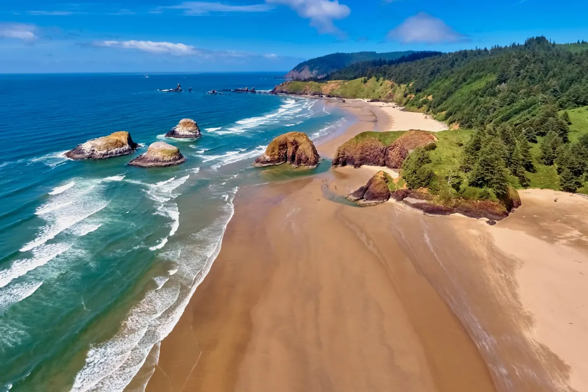 An exterior perspective of a sandy beach bordered by striking rock formations, rolling ocean waves, and green hills in the distance. The image exudes serenity and the inviting allure of outdoor exploration.