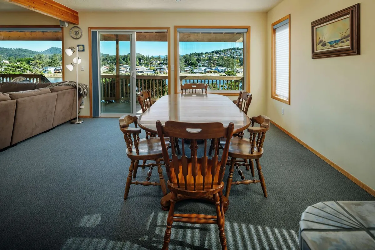 A dining area with a large table sits beside broad windows, offering views of the surrounding landscape. The proximity to the living area and sofa highlights the open-concept layout, ideal for group meals and shared moments.