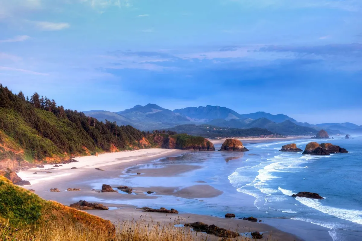 A sweeping landscape view of a sandy beach with dramatic rock formations, gentle waves lapping at the shore, and distant mountains creating an awe-inspiring natural backdrop. The tranquil scene highlights the vastness of nature and the sense of peaceful escape.