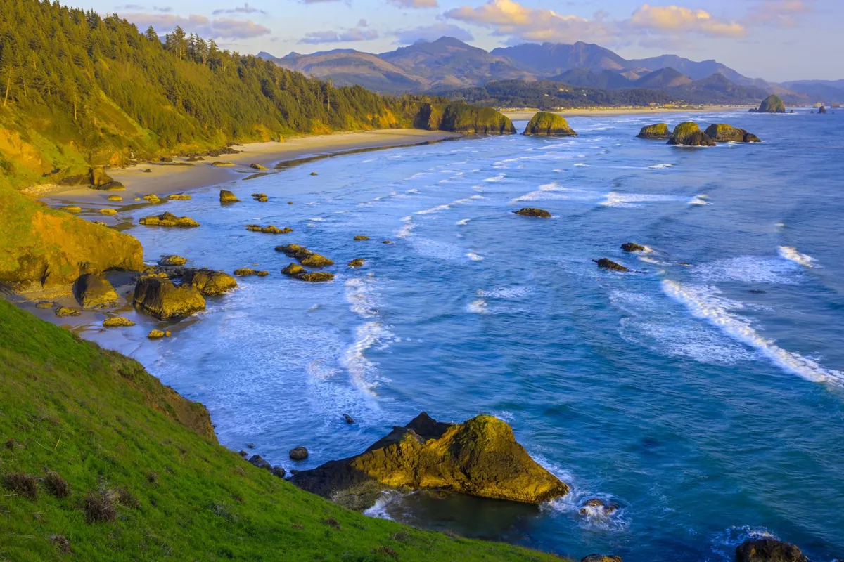 Expansive ocean waves roll onto a broad sandy beach, framed by striking rock formations and a forested hillside, with distant mountains on the horizon. The scene is filled with possibilities for outdoor adventure and relaxation.