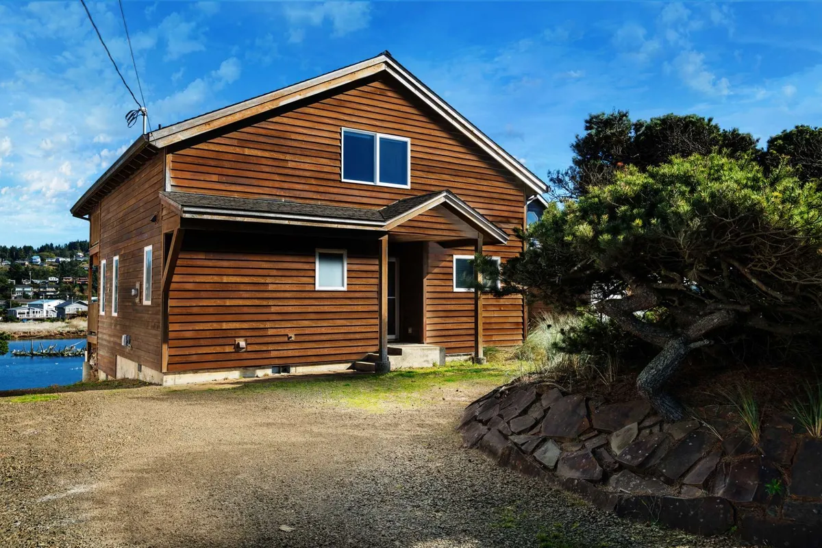 A wide exterior shot showing the wooden facade of the house with large windows, surrounded by a landscaped garden. The inviting porch overlooks the water, suggesting a seamless transition from indoor comfort to the outdoor riverfront space described in the section.