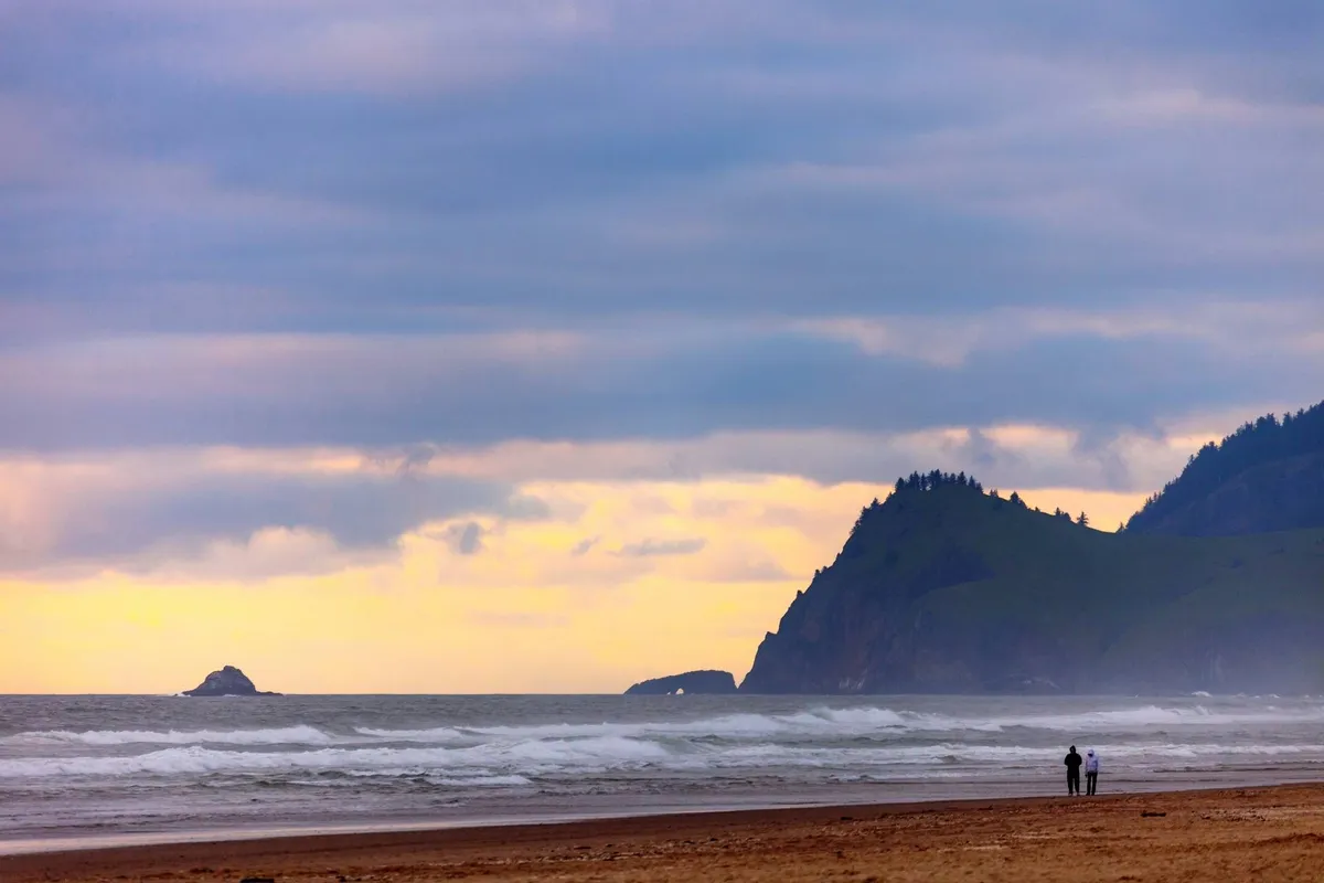 A couple strolls along the shoreline at sunset, with distant cliffs and the endless ocean creating a romantic, tranquil atmosphere. The image evokes a sense of escape and connection to nature.