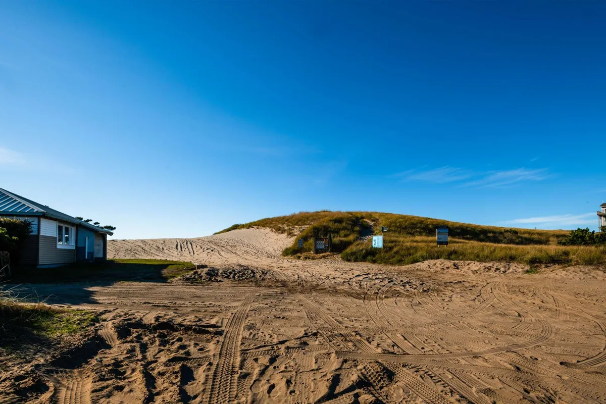 A sandy beach path winds through gentle dunes, leading toward the ocean. Nearby structures hint at a welcoming, walkable neighborhood just steps from the sand. The warm sunset glow invites exploration and relaxation.