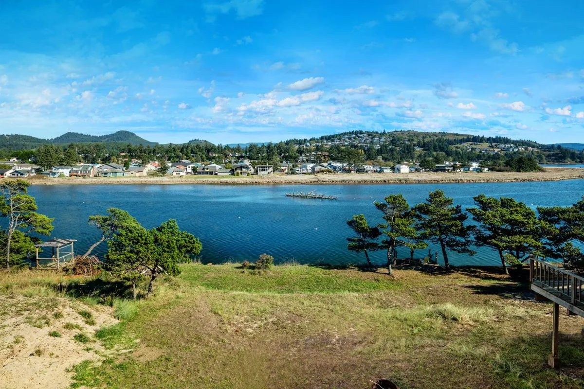 The image showcases a peaceful riverfront scene with tranquil water, tree-lined banks in the foreground, distant rolling hills, and the quaint town visible across the water. The setting is serene, inviting, and perfectly captures the unique riverside charm of Pacific City, with opportunities for sunset watching, wildlife spotting, and easy access to community activities by the shore.