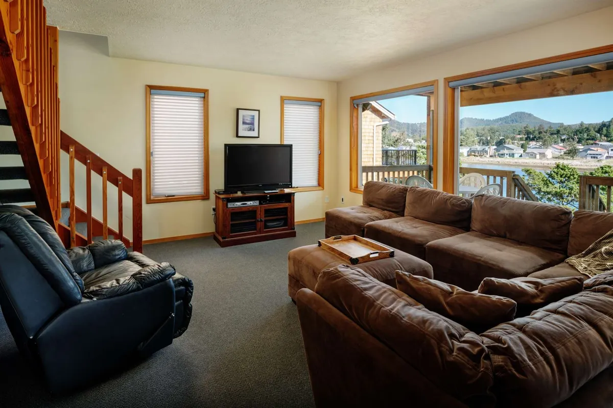 This image displays a spacious living room featuring a large sectional sofa positioned to face the home's only TV. Expansive windows frame panoramic river views, bathing the space in natural light and creating an inviting atmosphere for relaxation, socializing, and movie nights.